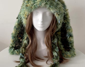 May include: A green and yellow faux fur hat with a hood and long braided tassels. The hat is made of a shaggy, textured material and is displayed on a white mannequin head. The hat has a whimsical, forest-like appearance.