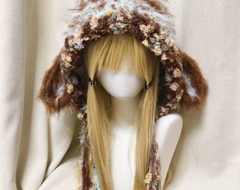 May include: A fuzzy, hooded hat featuring a blend of brown, white, and light blue yarn. The hood is trimmed with textured, multi-colored fringe in shades of brown, cream, and blue, and includes long, dangling braided ties on each side.