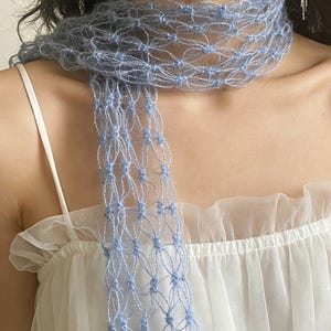 Peut inclure: Une &eacute;charpe bleu clair &agrave; tissage ouvert. L'&eacute;charpe est drap&eacute;e autour du cou et pend devant. Le design d&eacute;licat et a&eacute;r&eacute; est fait d'une mati&egrave;re douce et duveteuse. L'&eacute;charpe est port&eacute;e sur un haut blanc.
