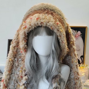 May include: A fluffy, hooded hat in shades of brown, beige, and orange, with colorful yarn accents. The hat has long, braided tassels. This unique headwear is perfect for cosplay or adding a whimsical touch to any outfit.