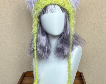 May include: A fuzzy, multi-colored knit hat featuring pointed cat ears, a lavender upper section, and a bright lime green lower band. Two long, lime green braided earflaps extend downward, each ending in a star-shaped pom-pom detail.