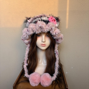 May include: A fuzzy, light pink and multi-colored textured hat featuring two cat-like ears on top, a pink paw print patch, and two long side flaps ending in large, round, soft pink pom-poms. The hat has a shaggy, yarn-like appearance throughout.