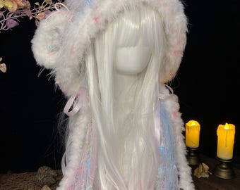 May include: A fluffy, hooded accessory featuring a white, blue, and pink textured yarn blend. The hood includes two round, soft pink pom-poms hanging from long, ribbon-accented ties. The material has a shaggy, faux-fur appearance with pastel color flecks throughout.