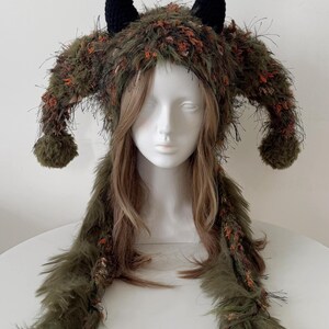 May include: A textured olive green hat featuring long side flaps, two large side protrusions with pom-poms at the ends, and two small black crocheted horns on top. The hat is crafted from a shaggy, multi-tonal yarn with flecks of orange throughout.