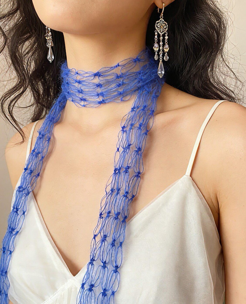 Peut inclure: Gros plan sur une &eacute;charpe bleue et des boucles d'oreilles. L'&eacute;charpe est faite d'une mati&egrave;re d&eacute;licate en forme de filet et se drape autour du cou. Les boucles d'oreilles sont argent&eacute;es avec des accents en forme de cristal. Le fond est de couleur neutre.