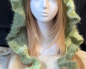 Peut inclure: Une capuche en tricot duveteux vert clair avec un design à bords volantés. La capuche présente une construction en fil doux et texturé dans une teinte vert sauge pâle, avec de longs panneaux volantés fluides tombant sur les côtés.
