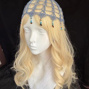May include: A light blue crocheted head covering with a delicate net design. The headpiece is adorned with dangling beads in shades of blue, brown, and white. The head covering is displayed on a mannequin head with long, wavy blonde hair.