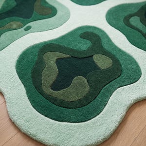 Pu&ograve; includere: Un tappeto tufted caratterizzato da un motivo organico astratto con forme irregolari stratificate in varie tonalit&agrave; di verde, tra cui verde bosco scuro, oliva e menta chiaro, su uno sfondo bianco sporco con un pelo testurizzato.