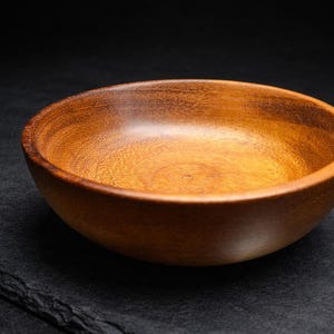 Puede incluir: Un cuenco de madera hecho a mano con un acabado natural y cálido. El cuenco tiene una forma lisa y redondeada y un color marrón intenso. El cuenco de madera es perfecto para servir ensaladas, aperitivos o como pieza decorativa.