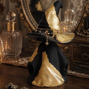 May include: A black witch hat sculpture with gold leaf accents, featuring a draped base and a brim adorned with a gold-toned chain necklace. The chain includes small star, moon, and bead charms that hang from the hat's brim in a decorative arrangement.