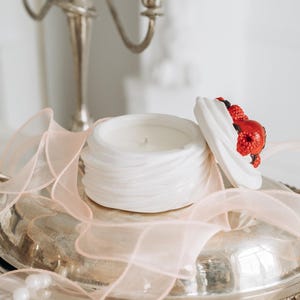 Pode incluir: Uma vela de cerâmica branca em um recipiente decorativo com uma tampa adornada com bagas vermelhas e pretas. A vela está em uma bandeja de prata com uma fita rosa, ao lado de um castiçal de prata. Um colar de pérolas também é visível.