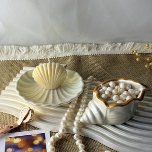 Op de afbeelding: Een crèmekleurige kaars in de vorm van een schelp op een bijpassend schaaltje, een schelpvormige kom gevuld met kleine witte bolletjes, een snoer witte parels, een roségouden lontknipper en een foto met de tekst "Бережіть себе".