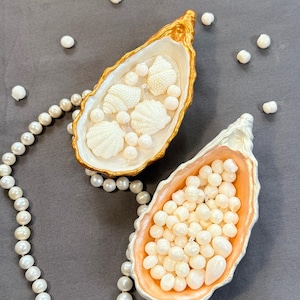 May include: Two decorative oyster shells used as trinket dishes. One shell features a gold-painted rim and contains small white seashell-shaped ornaments. The other shell has a peach-coloured interior filled with numerous loose white pearls.