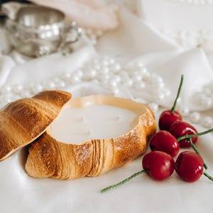 Puede incluir: Una vela con forma de croissant con un interior de cera blanco cremoso y una parte superior separada. La vela se encuentra sobre una superficie de satén blanco, junto a un grupo de cerezas rojas. Un juego de té plateado y perlas están en el fondo.