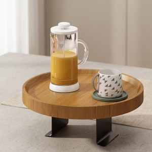 May include: A wooden serving tray with a clear glass French press filled with orange liquid, a patterned mug, and a coaster. The tray has a round shape and is supported by black metal legs.