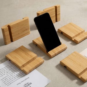 May include: A collection of bamboo phone stands. One stand holds a black smartphone. The stands are rectangular with a slot to hold a phone. The stands are made of light-colored bamboo.