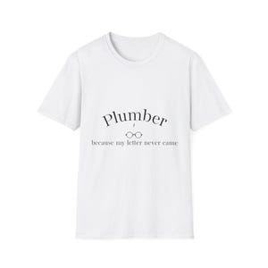 Peut inclure: T-shirt blanc avec le mot "Plumber" en écriture noire, avec un petit dessin de lunettes, et la phrase "because my letter never came". Le t-shirt a un col rond et des manches courtes.