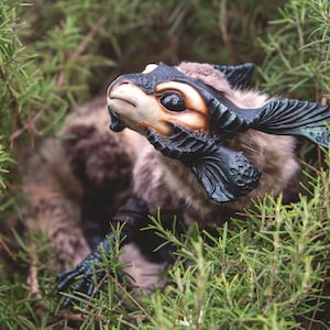 Peut inclure: Une créature en peluche détaillée avec un visage beige, de grands yeux noirs et des accents bleu foncé. La créature a un corps poilu dans les tons de gris et de marron, nichée dans un feuillage vert.
