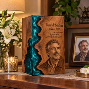 Op de afbeelding: Een rechthoekige houten urn met een verticale inleg van wervelende blauwgroene hars. De voorzijde is gegraveerd met de naam David Miller, de jaren 1960-2023, een herdenkingscitaat en een portret van een man met een baard.