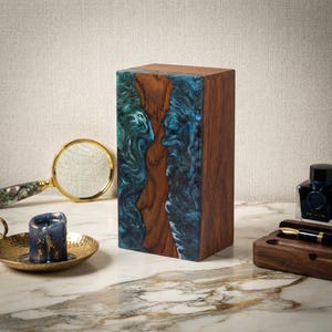 May include: A rectangular block made of dark brown wood and swirling blue and teal resin. The resin features a marbled, wave-like pattern that contrasts with the natural wood grain. The object stands upright on a flat surface.