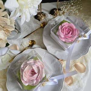 May include: Two pink rose-shaped desserts in clear boxes, presented on white plates with decorative edges. Each box is tied with a white ribbon and rests on a green leaf. The scene includes gold spoons, white flowers, and pearl accents.