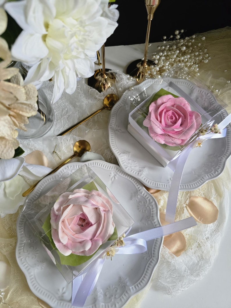 May include: Two pink rose-shaped desserts in clear boxes, presented on white plates with decorative edges. Each box is tied with a white ribbon and rests on a green leaf. The scene includes gold spoons, white flowers, and pearl accents.