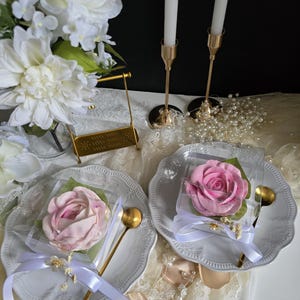 May include: Two white plates with a pink rose-shaped treat in clear packaging, tied with white ribbon and a gold spoon. Gold candle holders with white candles and a bouquet of white flowers are in the background.