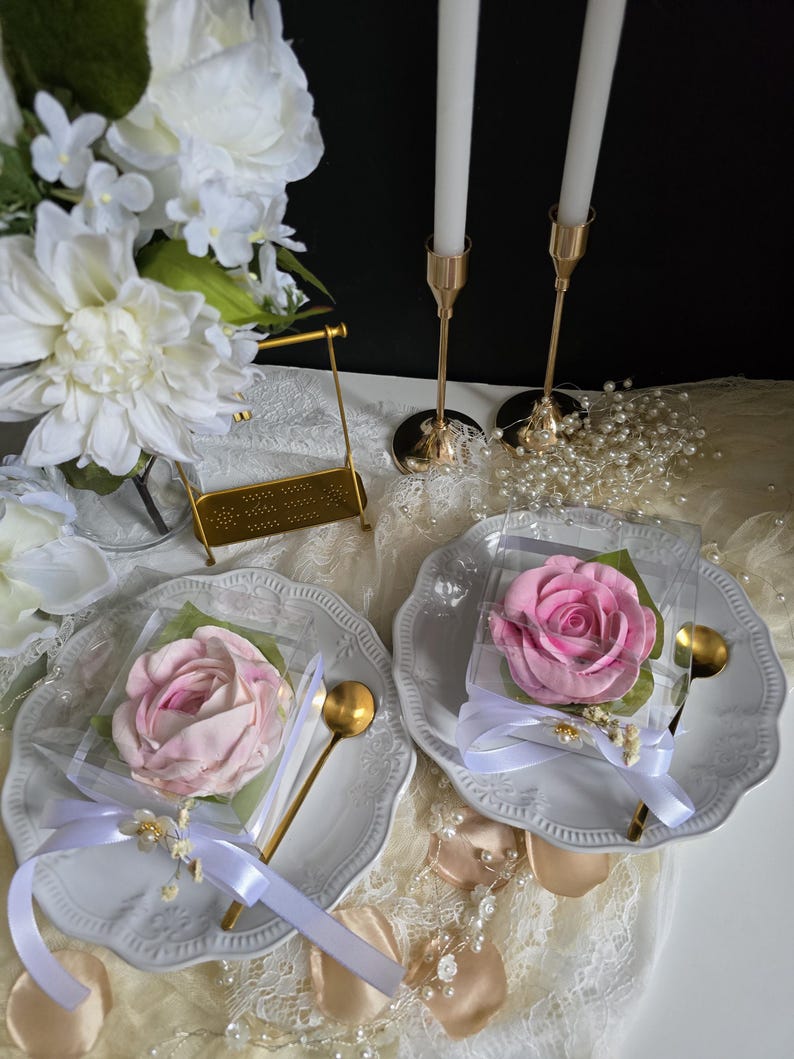 May include: Two white plates with a pink rose-shaped treat in clear packaging, tied with white ribbon and a gold spoon. Gold candle holders with white candles and a bouquet of white flowers are in the background.