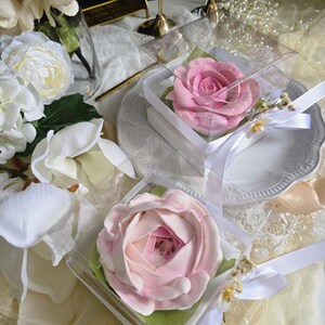 May include: Two pink rose-shaped items in clear boxes with white ribbons. The roses are on green paper and sit on white plates. The background includes white flowers, pearls, and decorative elements.