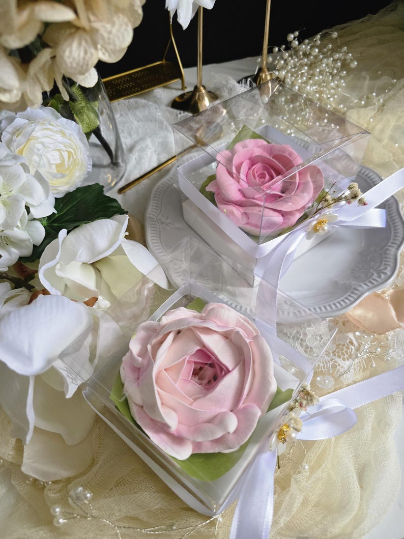 May include: Two pink rose-shaped items in clear boxes with white ribbons. The roses are on green paper and sit on white plates. The background includes white flowers, pearls, and decorative elements.