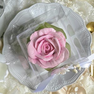 May include: A pink rose-shaped confection, presented in a clear square box, rests on a white, ornate plate. The dessert is surrounded by green leaves and a white ribbon. The background includes lace and pearl accents.