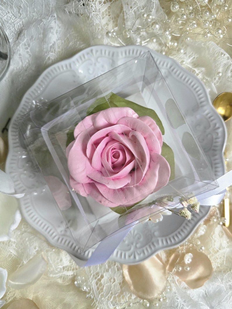 May include: A pink rose-shaped confection, presented in a clear square box, rests on a white, ornate plate. The dessert is surrounded by green leaves and a white ribbon. The background includes lace and pearl accents.
