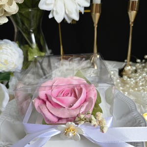 May include: A pink rose in a clear, square box, tied with a white ribbon and a decorative flower. The rose is the focal point, with green leaves and small white flowers. The background includes white flowers, candles, and pearls.