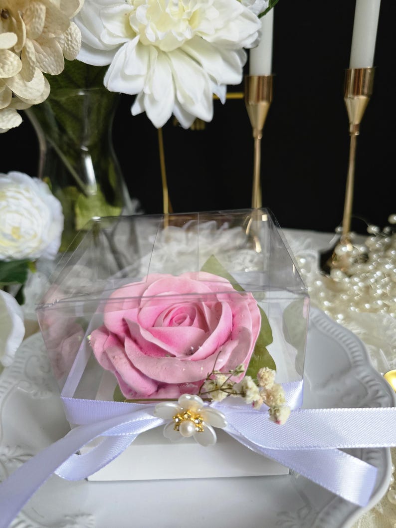 May include: A pink rose in a clear, square box, tied with a white ribbon and a decorative flower. The rose is the focal point, with green leaves and small white flowers. The background includes white flowers, candles, and pearls.