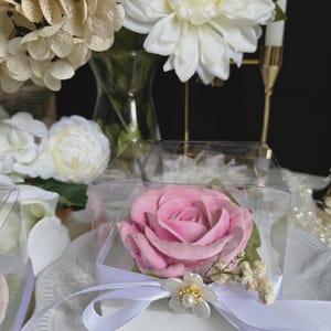 May include: A pink rose in a clear box tied with a white ribbon and a decorative flower. The rose is the focal point, set against a backdrop of white and cream-colored flowers and a white plate.