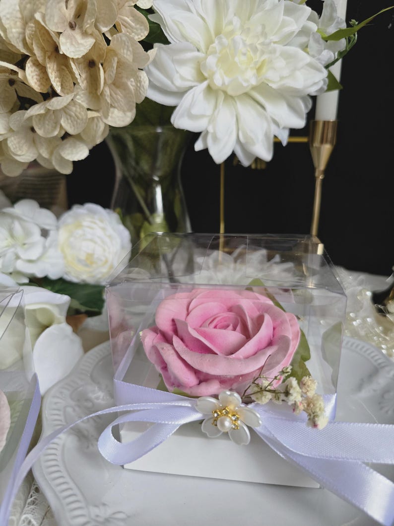 May include: A pink rose in a clear box tied with a white ribbon and a decorative flower. The rose is the focal point, set against a backdrop of white and cream-colored flowers and a white plate.