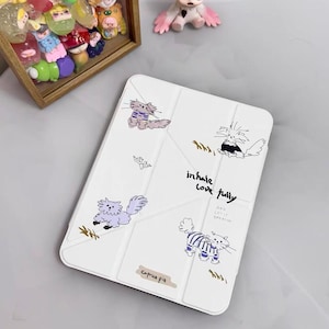 May include: White tablet case with cartoon cat illustrations in blue and black. The case has the text "inhale love fully" and "caprice pie". A wooden box with colorful figurines and a pink doll are also in the image.