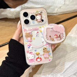 以下が含まれることがあります： ピンクのハローキティのデザインが施された透明な携帯電話ケース。ケースには、複数のハローキティのキャラクター、食べ物、そして「cute」という言葉が描かれています。ハローキティのデザインが施されたお揃いのポップソケットも表示されています。