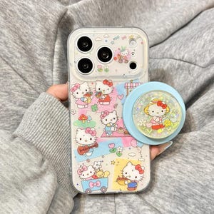 Puede incluir: Funda transparente para tel&eacute;fono con ilustraciones de Hello Kitty y un soporte popsocket azul claro. La funda tiene un fondo transparente con im&aacute;genes coloridas de Hello Kitty. El soporte popsocket tiene una base azul y una parte superior transparente con un dise&ntilde;o de Hello Kitty.