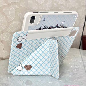 May include: A white phone case with a blue and white plaid pattern and cartoon illustrations of a bunny and a dog. The phone case is propped up, revealing the phone's camera and a matching design on the back. The text "YQQXCase" is visible.