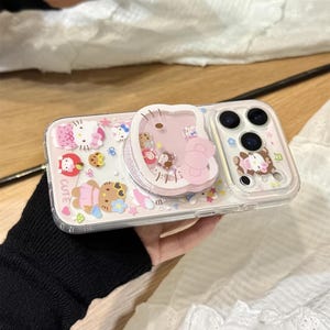 以下が含まれることがあります： ピンクと白のハローキティのデザインの透明な携帯電話ケース。ケースには、ピンクのハローキティのデザインのポップアップグリップと、クマのデザインの透明な部分があります。「CUTE」という文字がケースに表示されています。