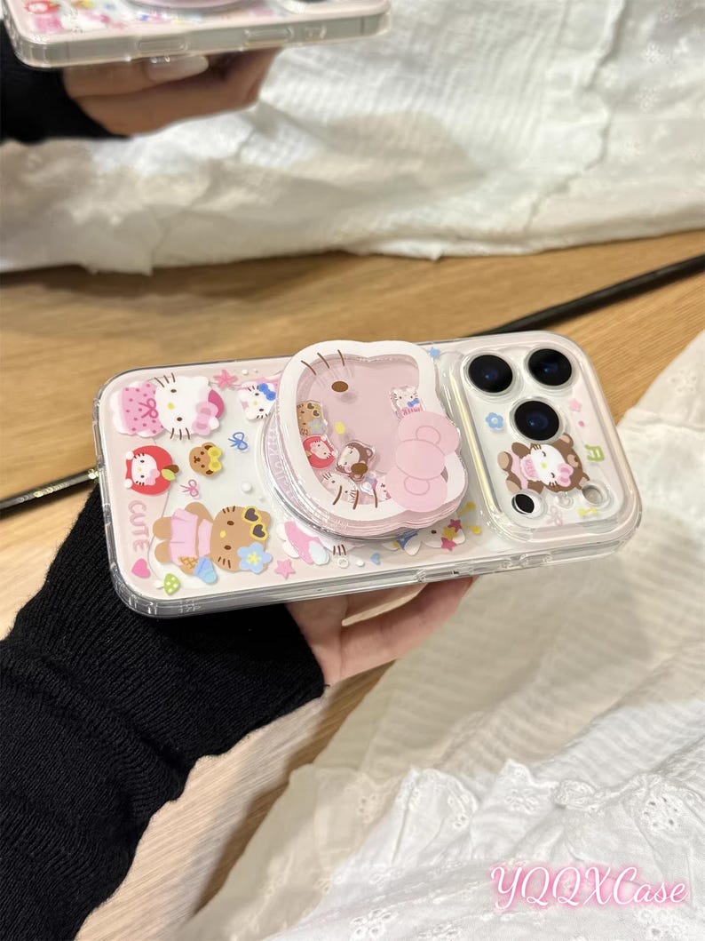 以下が含まれることがあります： ピンクと白のハローキティのデザインの透明な携帯電話ケース。ケースには、ピンクのハローキティのデザインのポップアップグリップと、クマのデザインの透明な部分があります。「CUTE」という文字がケースに表示されています。