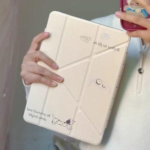 May include: Cream-colored tablet case with a geometric design and a cartoon dachshund illustration. The case features the text "Refuse to let it get to you" and "Be yourself, bright smile". The case is held in a person's hands.