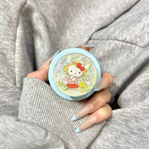 Puede incluir: Un soporte redondo para tel&eacute;fono de color azul claro con un dise&ntilde;o de Hello Kitty. El dise&ntilde;o incluye a Hello Kitty con un sombrero y rodeada de flores. El soporte lo sostiene una persona con una sudadera gris.