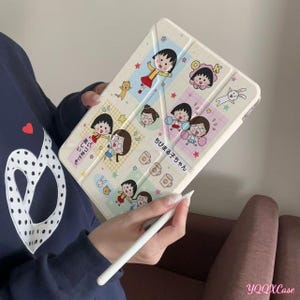 May include: A beige tablet case featuring a colorful cartoon design with multiple panels. Each panel displays a different illustration of a girl with black hair, along with Japanese text and other graphics. A white stylus is held near the case.