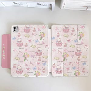 May include: Pink iPad case with a repeating pattern of a cartoon cat, cupcakes, bows, and flowers. The case is open, revealing the interior design. The text "Sweet kitty" is repeated throughout the design.