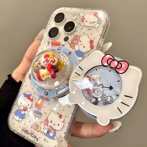 May include: Clear phone case featuring Hello Kitty designs and a pop-up bubble with miniature figures. The case has a white Hello Kitty-shaped clock with a pink bow and a rotating liquid-filled charm. The text "YQQXCase" is visible.