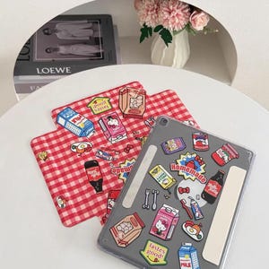 May include: A clear tablet case with various food-themed stickers, including a Hello Kitty milk carton and a bottle of soda. Two red and white checkered placemats are also visible, along with a vase of pink flowers.