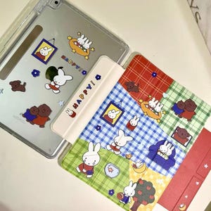 May include: Clear tablet case and colorful covers featuring cartoon bunnies and bears. The covers have a patchwork design with blue, green, and red sections. The word "HAPPY!" is printed on one cover. The tablet case has stickers of the same characters.