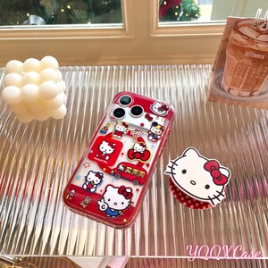 May include: A red and clear phone case with Hello Kitty designs. The case has a red border and a clear back, displaying various Hello Kitty illustrations and a red school bus. A Hello Kitty phone grip is also visible.